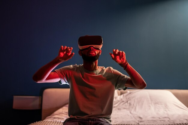 젊은 아프리카계 미국인이 은 네온 조명 아래 침대에 앉아 미래의 메타버스 VR 헤드을 사용하고 있습니다.