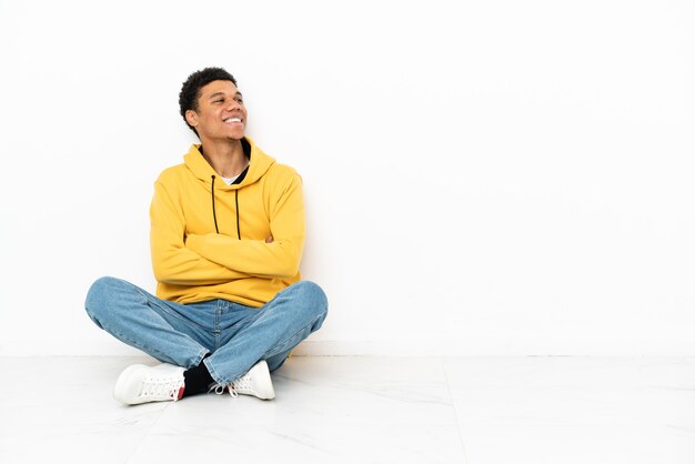 Giovane uomo afroamericano seduto sul pavimento isolato su sfondo bianco felice e sorridente