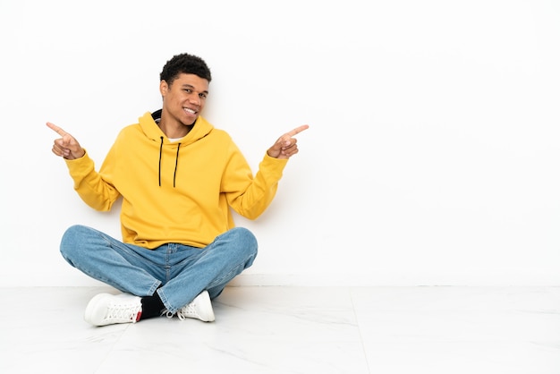 Young African American man sitting on the floor isolated on white background pointing finger to the laterals and happy