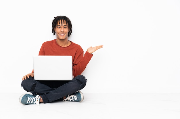 Young african american man sitting on the floor and working with his laptop holding copyspace imaginary on the palm to insert an ad