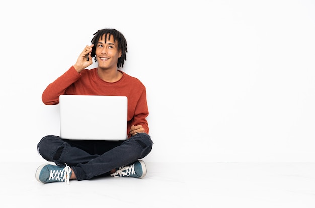 Young african american man sitting on the floor and working with his laptop keeping a conversation with the mobile phone