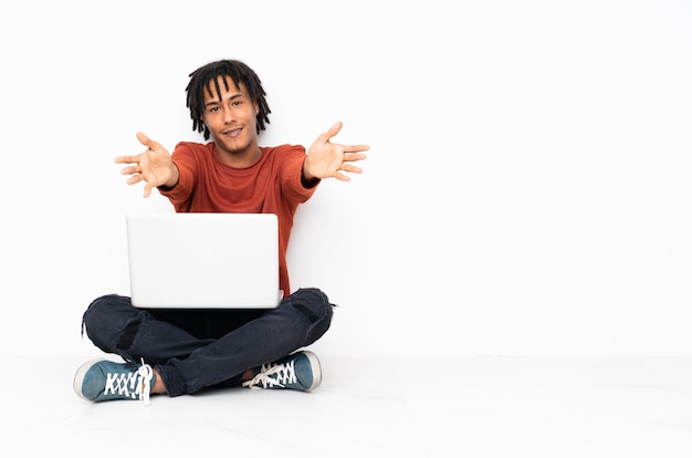 Молодой афроамериканец человек сидит на полу и работает со своим ноутбуком, представляя и приглашая прийти с рукой