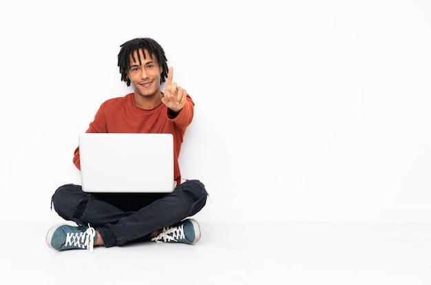 Giovane uomo afroamericano che si siede sul pavimento e che lavora con il suo computer portatile che mostra e che solleva un dito
