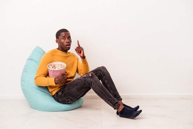 흰색 배경에 격리된 팝콘을 먹고 있는 퍼프에 앉아 있는 젊은 아프리카계 미국인 남자는 훌륭한 아이디어, 창의성 개념을 가지고 있습니다.