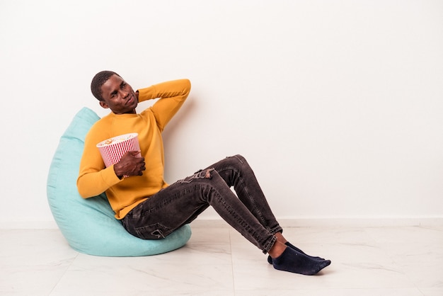 Young African American man sitting on a puff eating popcorn isolated on white background touching back of head, thinking and making a choice.