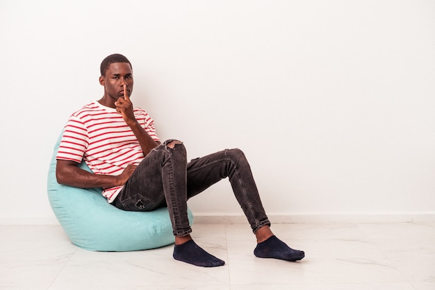 젊은 아프리카계 미국인 남자는 비밀을 유지하거나 침묵을 요구하는 흰색 배경에 고립 된 퍼프에 앉아 있습니다.