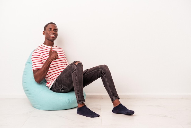 Young african american man sitting on a puff isolated on white background smiling and raising thumb up