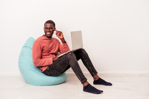 Young African American man sitting on a puff using laptop isolated on white background covering ears with hands.