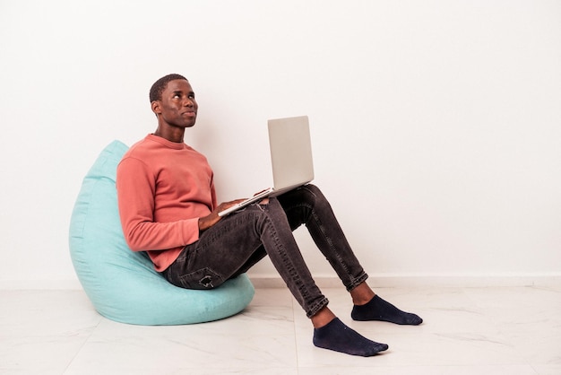 Young African American man sitting on a puff using laptop isolated on white background dreaming of achieving goals and purposes