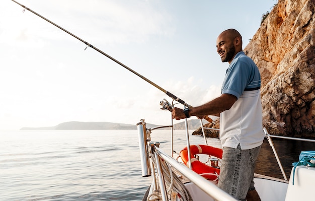 Young african american man standing with fishing rod on a sailboat fishing in open sea on sunset