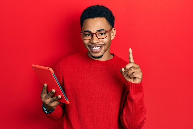 Young african american man using touchpad device surprised with an idea or question pointing finger with happy face, number one