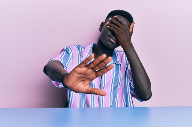 Young african american man wearing casual clothes sitting on the table covering eyes with hands and doing stop gesture with sad and fear expression embarrassed and negative concept