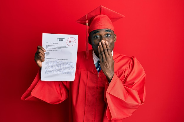 Young african american man wearing graduation cap and ceremony robe showing passed exam covering mouth with hand, shocked and afraid for mistake. surprised expression