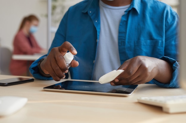 Foto giovane uomo afro-americano che pulisce tablet con salviettine igienizzanti mentre si lavora alla scrivania in ufficio post pandemia