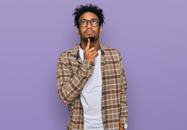 Young african american man with beard wearing casual clothes and glasses thinking concentrated about doubt with finger on chin and looking up wondering