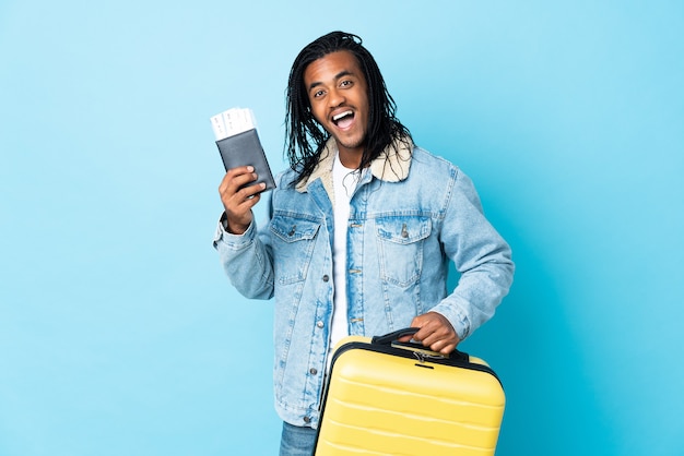 Young African American man with braids isolated on blue background in vacation with suitcase and passport