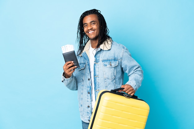 Young African American man with braids isolated on blue wall in vacation with suitcase and passport