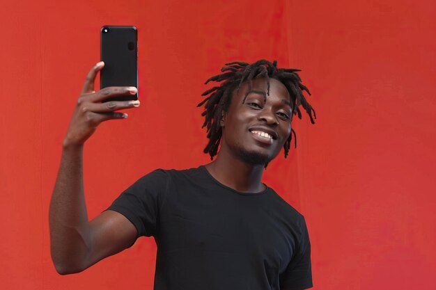 Young African American man with dreadlocks taking selfie with victory sign on red wall