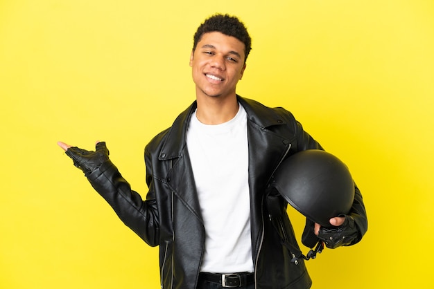 Giovane uomo afroamericano con un casco da motociclista isolato su sfondo giallo che estende le mani di lato per invitare a venire