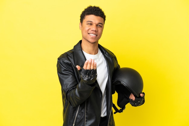 Young African American man with a motorcycle helmet isolated on yellow background inviting to come with hand. Happy that you came