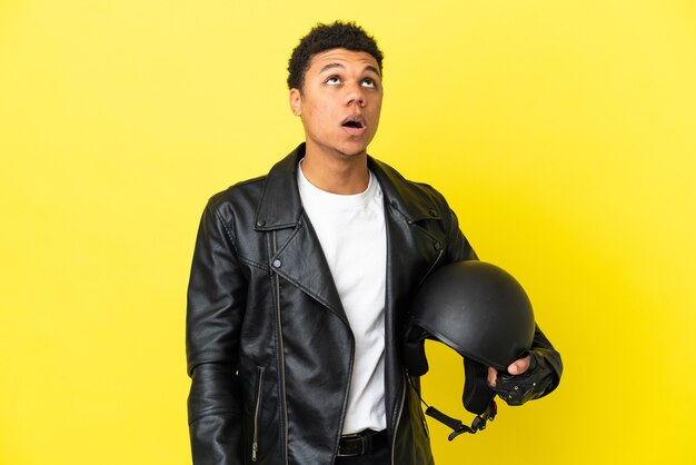 Young African American man with a motorcycle helmet isolated on yellow background looking up and with surprised expression