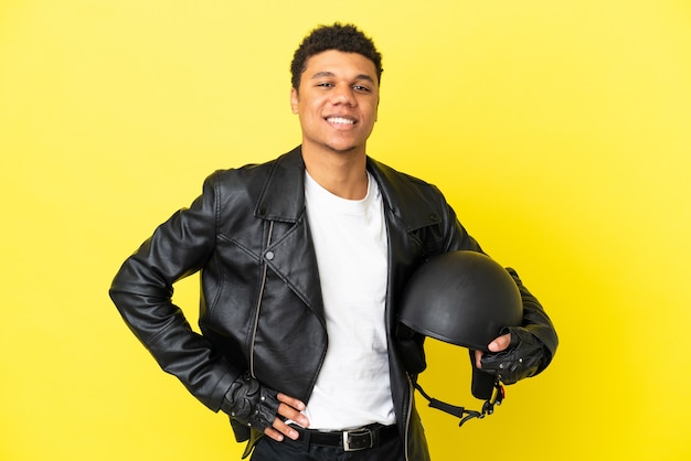 Молодой афро-американский мужчина с мотоциклетным шлемом, изолированные на желтом фоне, позирует с руками на бедрах и улыбается