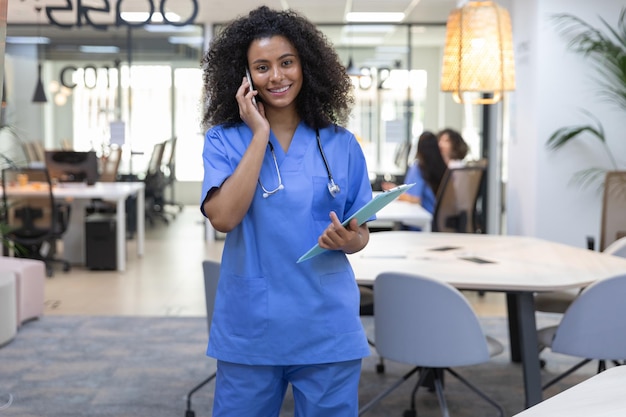 Giovane infermiera afroamericana che indossa l'uniforme blu parlando al telefono e guardando la fotocamera.