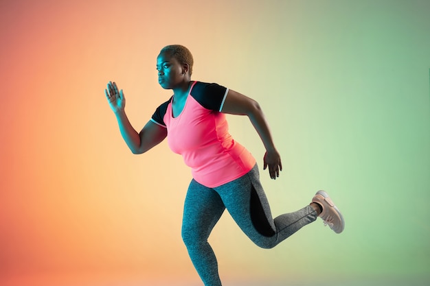 Young african-american plus size female model's training on gradient wall in neon light.