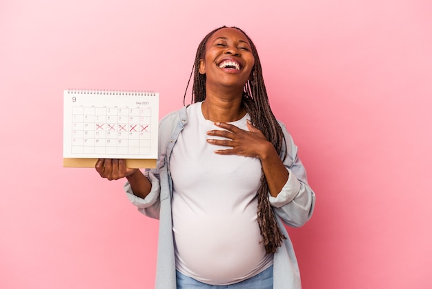 Young african american pregnant woman holding calendar isolated on pink background laughs out loudly keeping hand on chest.