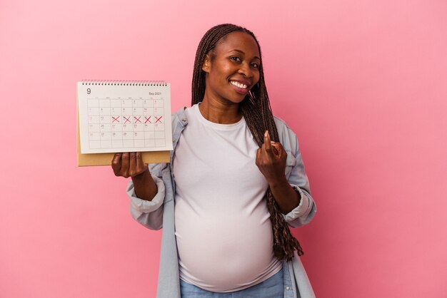 Молодая афро-американская беременная женщина, держащая календарь на розовом фоне, указывая пальцем на вас, как будто приглашая подойти ближе.