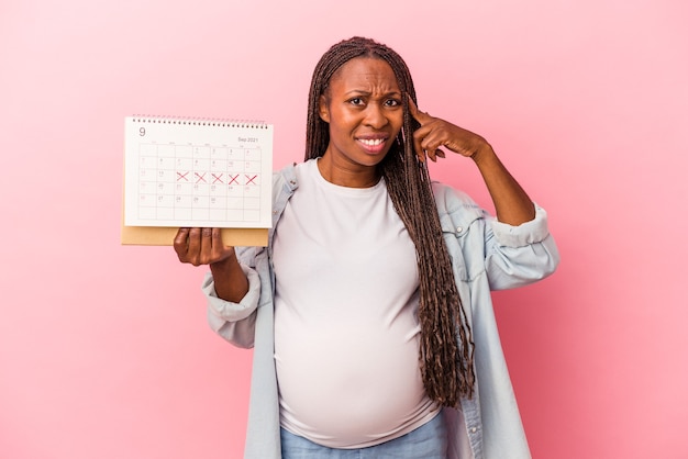 Giovane donna incinta afroamericana che tiene il calendario isolato su sfondo rosa che mostra un gesto di delusione con l'indice.