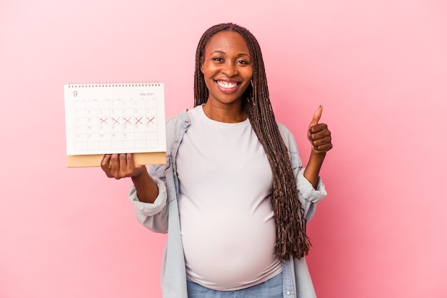 Young african american pregnant woman holding calendar isolated on pink background smiling and raising thumb up