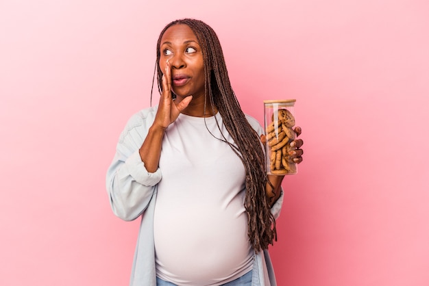 Young african american pregnant woman holding cookies isolated on pink background is saying a secret hot braking news and looking aside