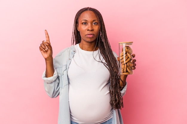 Young african american pregnant woman holding cookies isolated on pink background showing number one with finger