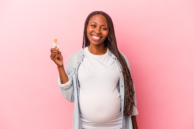 Giovane donna incinta afroamericana che tiene il ciuccio isolato su sfondo rosa felice, sorridente e allegro.