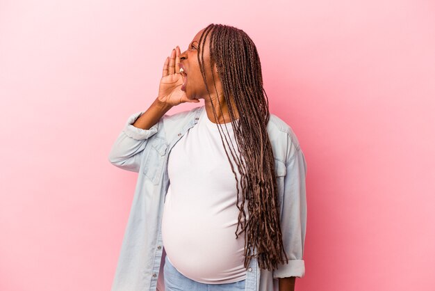 Giovane donna incinta afroamericana isolata su fondo rosa che grida e che tiene il palmo vicino alla bocca aperta.