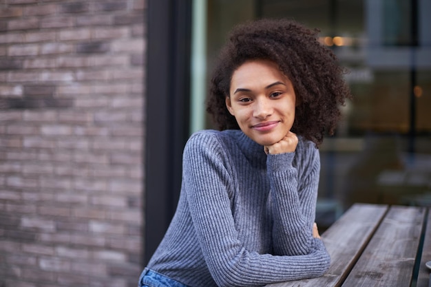 Young African American pretty woman smiling at camera sitting at a wooden table outdoors