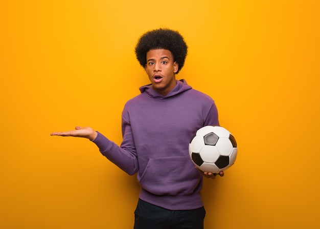 Young african american sport man holding a soccer ball holding something on palm hand