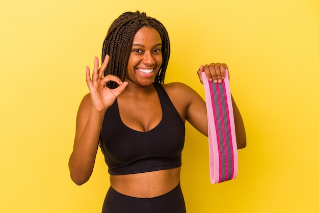 Young african american sport woman holding a elastic band isolated on yellow background  cheerful and confident showing ok gesture.