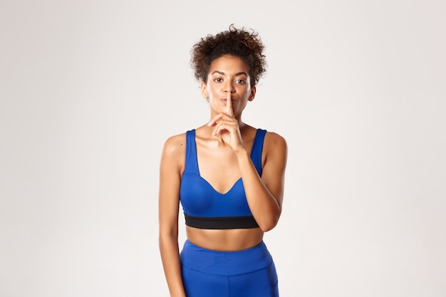 Young african-american sporty girl in blue sport outfit, asking keep voice down, shushing at camera