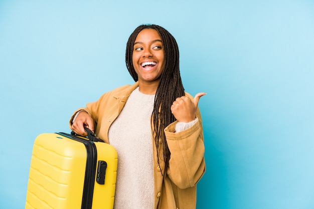 Young african american traveler woman holding a suitcase isolated points with thumb finger away, laughing and carefree.