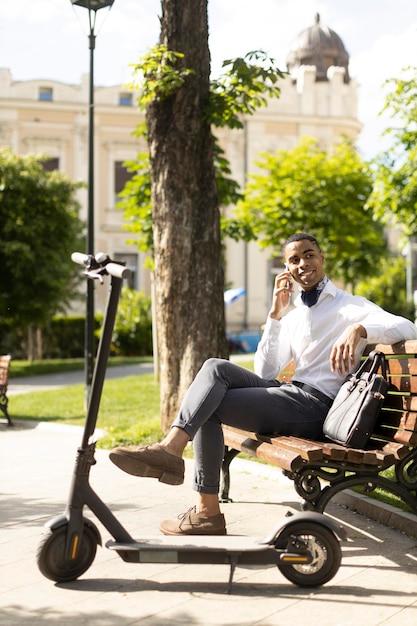 Young African American using mobile phone while sitting on the bench by electric scooter