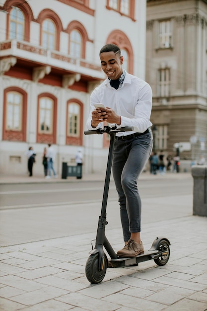 Young African American using mobile phone while standing with electric scooter on a street