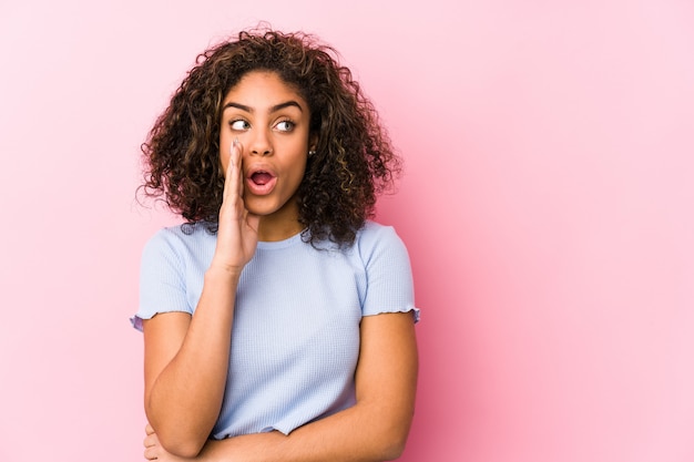 Young african american woman against a pink background is saying a secret hot braking news and looking aside