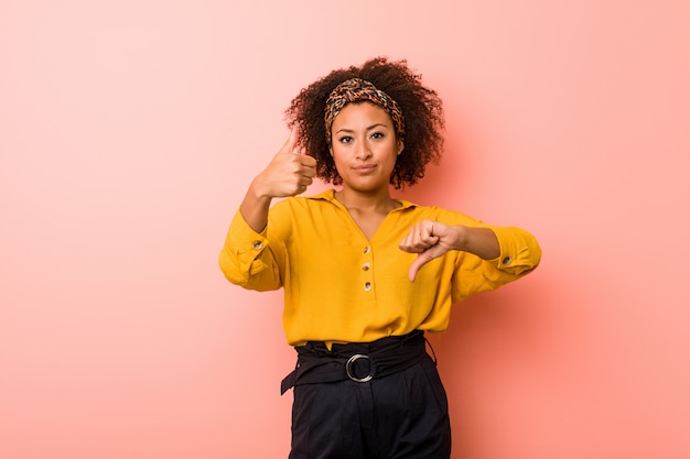 Photo young african american woman against a pink  showing thumbs up and thumbs down, difficult choose