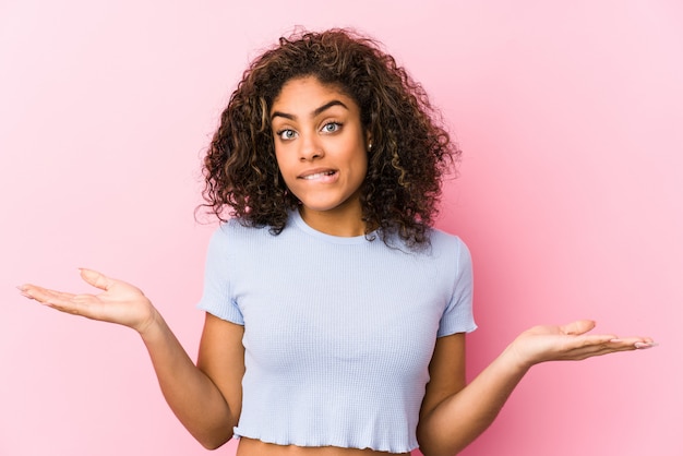 young african american woman against a pink wall confused and doubtful shrugging shoulders to hold a blank space.