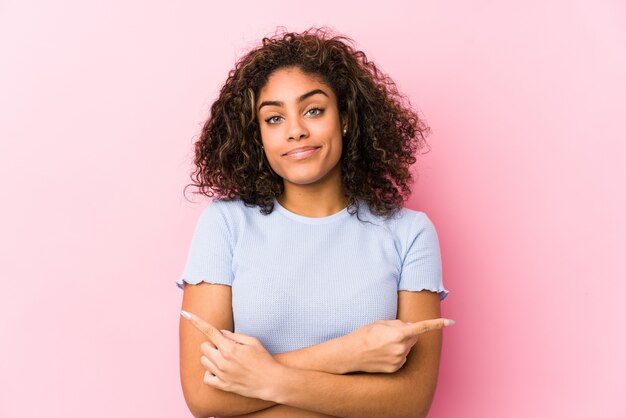 Young african american woman against a pink wall points sideways, is trying to choose between two options.