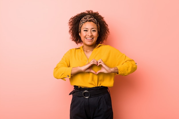 Young african american woman against a pink wall smiling and showing a heart shape with hands.