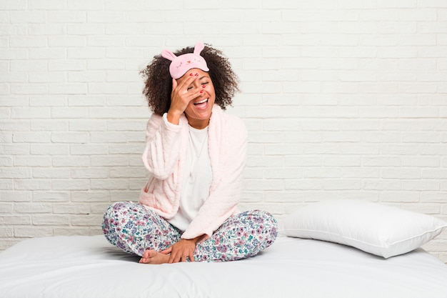 Young african american woman in the bed wearing pijama blink at through fingers, embarrassed covering face.