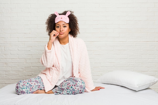 Young african american woman in the bed wearing pijama with fingers on lips keeping a secret.